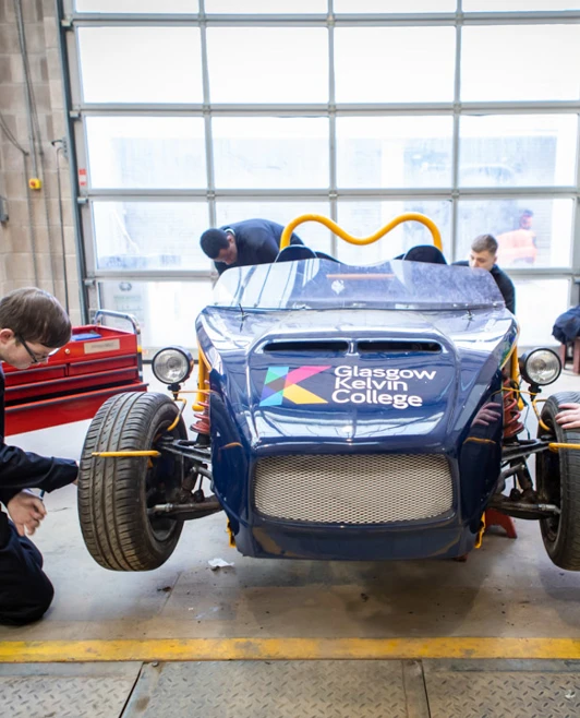Students in a workshop working on a blue Glasgow Kelvin College-branded kit car, focusing on different repair areas. Students in a workshop working on a blue Glasgow Kelvin College-branded kit car, focusing on different repair areas.